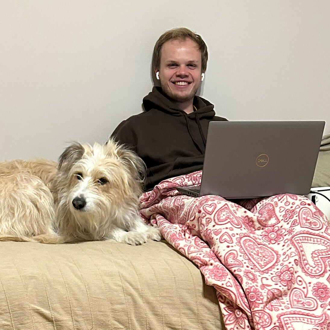 Dawid A man sitting on the bed with his laptop. He has earphones and blanket covering his legs. Big white dog is lying next to him.