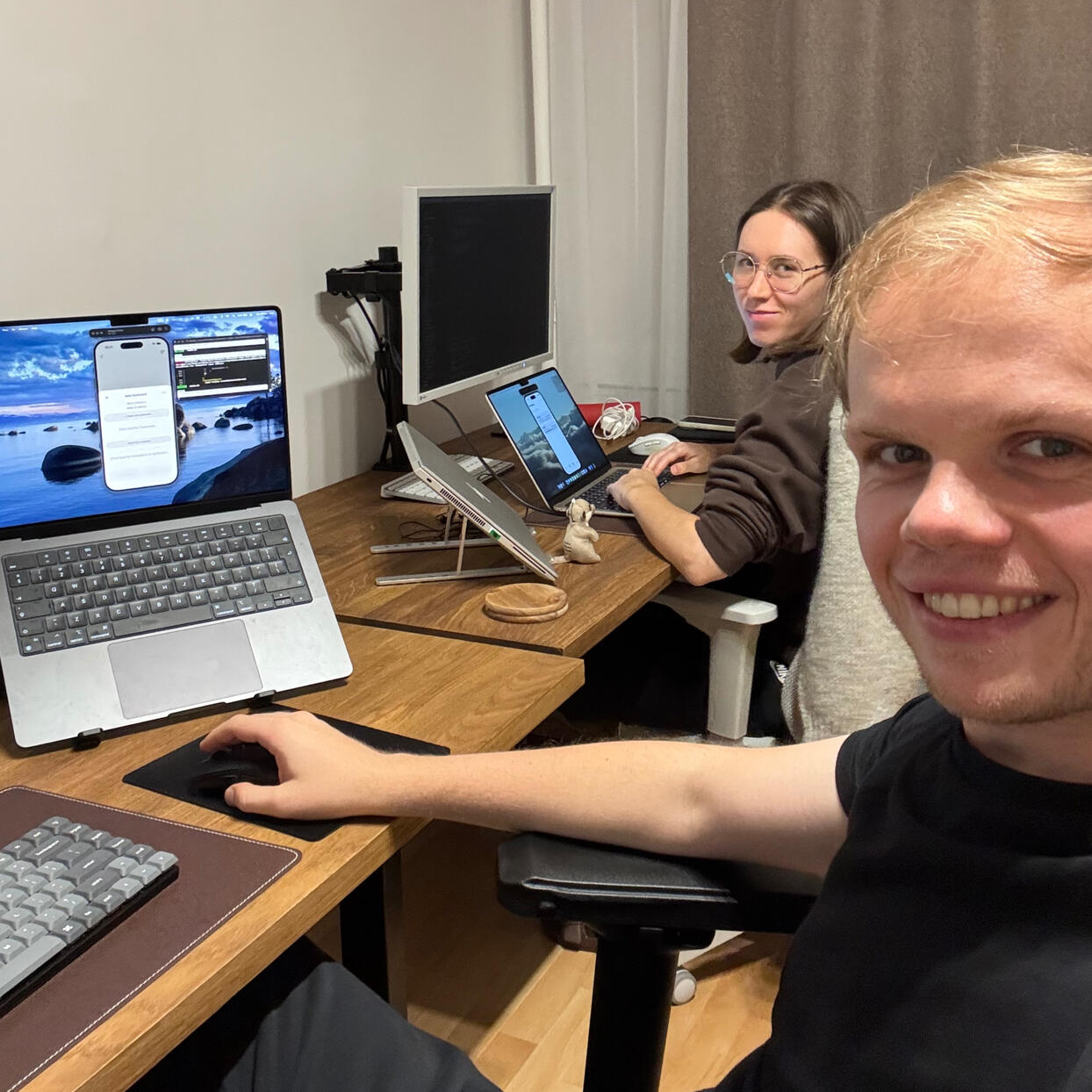 Dawid and Asia Foreground of the image shows a man sitting at the desk with his laptop on. In the background, a woman with glasses works on her laptop.